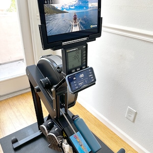 May include: Black Concept2 rowing machine with a tablet displaying a rowing video. The machine has a digital display showing workout metrics. A water bottle and phone are also visible. The machine is on a black mat.