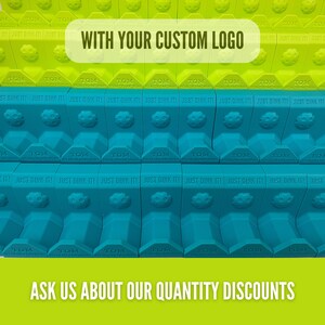Op de afbeelding: Rijen groene en blauwe pickleball-baanaccessoires met de tekst "WITH YOUR CUSTOM LOGO" en "ASK US ABOUT OUR QUANTITY DISCOUNTS". Elk accessoire heeft de tekst "TOM" en "JUST DINK IT!"