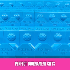 Op de afbeelding: Close-up van meerdere blauwe pickleball-items. Elk item heeft de tekst "PICKLEBALL FEST GRAND HAVEN" erop gedrukt. De tekst "PERFECT TOURNAMENT GIFTS" staat onderaan de afbeelding.
