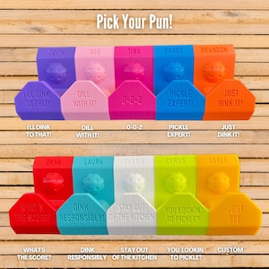 Op de afbeelding: Een verzameling kleurrijke pickleball-thema cadeaus met woordspelingen, waaronder "I'll Dink to That!" en "Just Dink It!". De items zijn in verschillende kleuren zoals paars, roze, blauw en oranje, met een pickleball-ontwerp. De tekst "Pick Your Pun!" staat bovenaan.