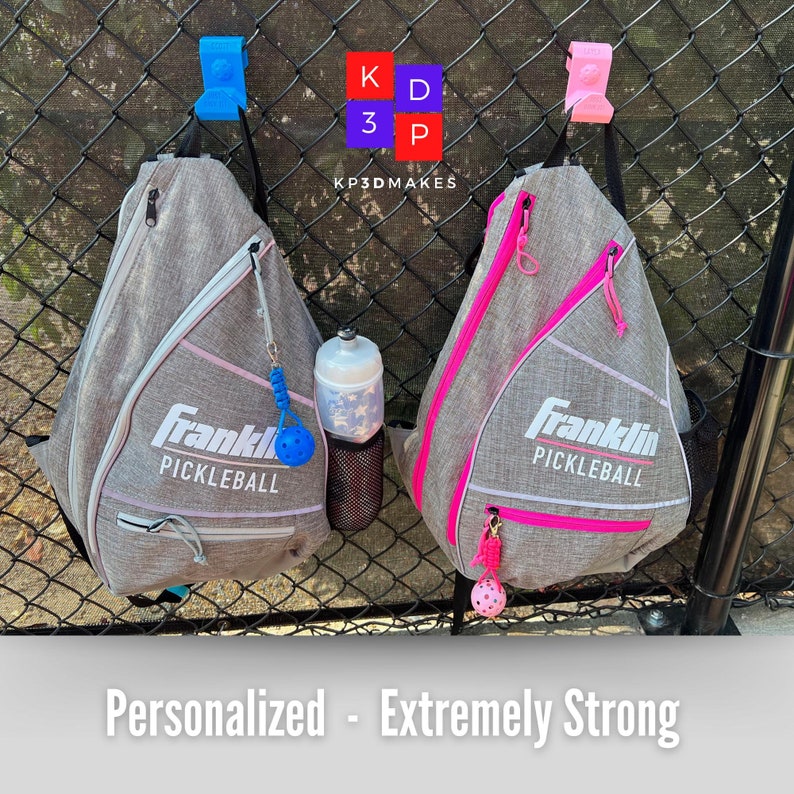 Op de afbeelding: Twee grijze Franklin Pickleball slingtassen met roze en witte accenten hangen aan een hek van gaas. Elke tas heeft een rits en een kleine bedel. Een waterfles en een blauwe en roze pickleball zijn ook zichtbaar. De tekst "Personalized - Extremely Strong" staat onderaan.