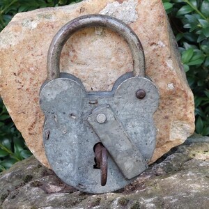 May include: A vintage, silver-colored padlock with a rusty hook. The padlock is sitting on a light brown rock.