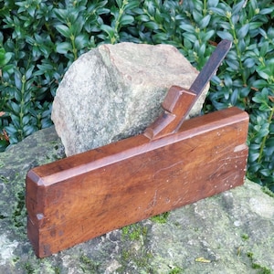 Op de afbeelding: Een vintage houten handvlak met een metalen mes. De vlak is rustend op een rots met groen gebladerte op de achtergrond.