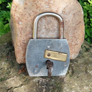 May include: A silver padlock with a brass keyhole and a brass plate that reads "SINCE" is secured to a large, flat, light brown rock.