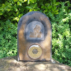 May include: A slate plaque with a carved image of a ship and a round, brass barometer. The plaque is mounted on a stone surface.