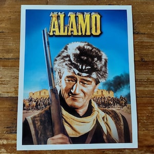 May include: A vintage movie poster for the film "The Alamo" featuring a close-up of John Wayne in a fur hat and holding a rifle. The poster has a blue background with the title "ALAMO" in yellow letters.