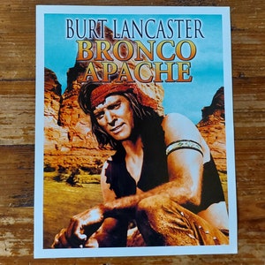 Peut inclure: Une affiche de film vintage mettant en scène Burt Lancaster dans le film "Bronco Apache". L'affiche a un fond bleu avec un schéma de couleurs rouge, blanc et bleu. Lancaster est représenté dans un décor rustique et extérieur, portant un bandeau et un gilet en cuir.