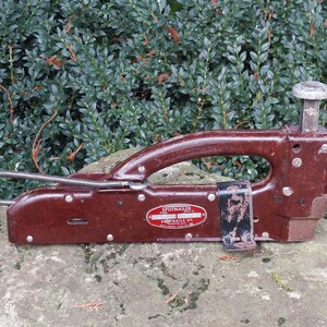 Op de afbeelding: Een bruine Spotnailer-spijkerpistool van metaal met een zwarte leren band en een zilveren metalen handvat. Het spijkerpistool ligt op een grijze steen.