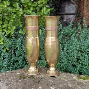 Puede incluir: Dos jarrones antiguos de latón dorado. Cada jarrón tiene una forma cilíndrica con una banda decorativa y una base acampanada. Los jarrones se exhiben sobre una superficie de piedra, con un fondo de follaje verde.