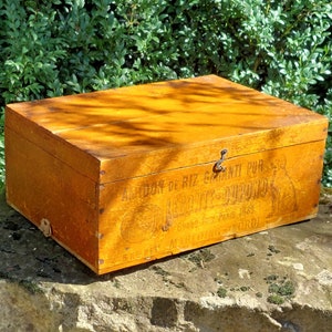 May include: A wooden box with a hinged lid and a metal latch. The box is labeled "A l'ordon de Riz Garanti Pur" and "La Petite-Dauphine" with a logo of a woman holding a bag of rice. The box is a vintage storage container.