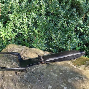 May include: A black metal garden tool with a long, curved handle and a sharp blade. The tool is resting on a rock with green foliage in the background.