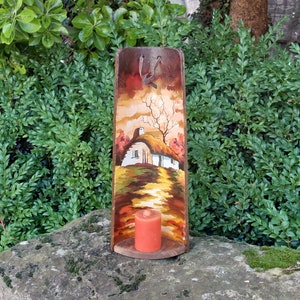 Può includere: Un portacandele in legno con una scena dipinta di un cottage in un paesaggio. Il portacandele ha un cinturino in pelle marrone e una piccola candela all'interno.