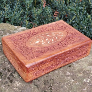 Peut inclure: Une boîte rectangulaire en bois avec des sculptures complexes et un motif floral. La boîte est de couleur brun clair et a une incrustation blanche au centre du couvercle.