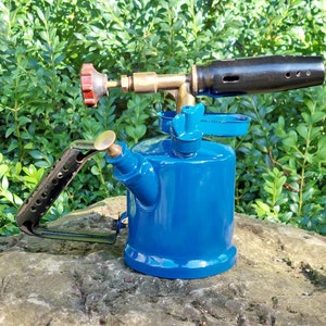 Op de afbeelding: Een blauwe metalen brander met een zwarte handgreep en een messing spuitmond. De brander heeft een zijhandgreep en een veiligheidsdop.