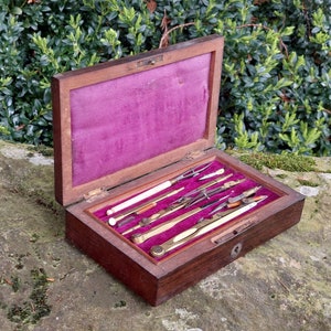 Può includere: Una scatola di legno con fodera in velluto, contenente un set di strumenti da disegno antichi, tra cui compassi, righelli e una matita. La scatola ha una serratura e un buco della serratura in ottone.