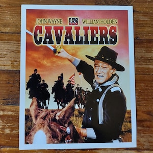 Peut inclure: Affiche de film vintage mettant en scène John Wayne et William Holden dans le film "Les Cavaliers". L'affiche a un fond de coucher de soleil rouge, orange et jaune avec une silhouette d'un groupe d'hommes à cheval. John Wayne est au premier plan, tenant une épée et regardant directement le spectateur.