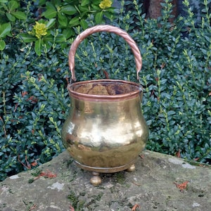 May include: A hammered brass bucket with a copper handle and three feet. The bucket is sitting on a stone surface.
