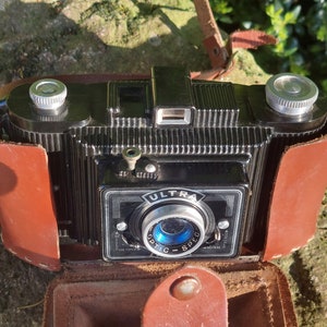 May include: A black vintage Ultra Optic Spec camera with a brown leather case. The camera has a silver lens and a silver knob on the top.
