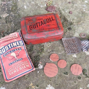 Peut inclure: Une boîte en métal rouge avec l'inscription "Necessaire Guttacoll Pour La Pneumatiques" et un paquet de rustines "Rustines Siamoises A TIRETTE" rouges et bleues. Le paquet indique "SANS DISSOLUTION SANS ESSENCE SANS RIEN".