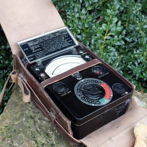 Könnte beinhalten: Vintage schwarzes elektrisches Testgerät mit rundem Zifferblatt und verschiedenen Messanzeigen, untergebracht in einer offenen braunen Ledertasche. Die Tasche zeigt das Zifferblatt des Instruments und eine Metallabdeckung mit Text. Das Gerät steht vor grünem Laub.