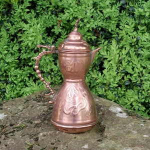 May include: A copper teapot with an ornate design and a curved handle. The teapot is sitting on a rock with green foliage in the background.