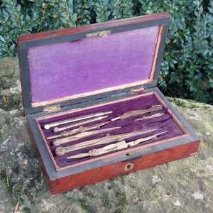 Può includere: Una scatola di legno con un rivestimento in velluto viola, contenente un set di compassi da disegno in ottone antico.