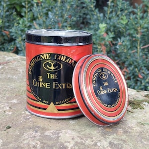 Op de afbeelding: Rode en zwarte blikken doos met gouden opschrift. De tekst luidt "Compagnie Coloniale The Chine Extra Selection des Meilleurs Thes".
