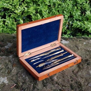 Può includere: Una scatola di legno con un rivestimento in velluto blu, aperta per rivelare un set di strumenti da disegno vintage in ottone. Gli strumenti includono compassi, righelli e un goniometro.