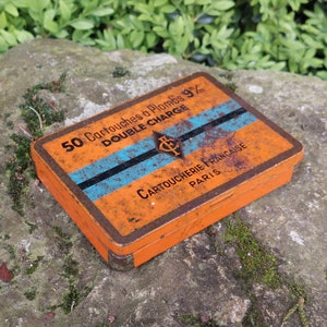 May include: A vintage orange and blue tin box with the text "50 Cartouches a Plombs 9/2m DOUBLE CHARGE CARTOUCHERIE FRANCAISE PARIS" printed on it.