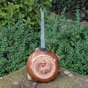 Puede incluir: Una sartén de color cobre con un mango largo plateado. La base de la sartén presenta un diseño en relieve de una persona con sombrero y abrigo. La sartén se encuentra sobre un fondo de follaje verde.