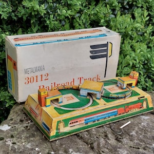 May include: A vintage tin toy train set with a red, white, and blue train, a green track, and yellow and red buildings. The set is in its original box, which is labeled "Metalmania 30112 Railroad Track".