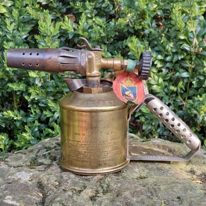 Op de afbeelding: Een vintage messing brander met een cilindrische brandstoftank en een mondstuk met meerdere gaten. De handgreep is geperforeerd en een label met tekst en een wapen is bevestigd. Gemaakt in Zweden.