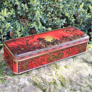 Könnte beinhalten: Eine rechteckige rote Dose mit einem dekorativen japanisch inspirierten Design. Der Deckel zeigt eine goldene Sonne, Bäume und einen Vogel. Die Seiten haben Blumen- und Blättermuster. Die Dose hat eine goldfarbene Umrandung.