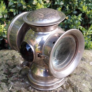 May include: Vintage silver carriage lamp with a clear glass lens and a black faceted jewel. The lamp has a domed top with embossed detailing and a circular base. The design includes a side lens and a small handle.