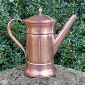 Puede incluir: Una tetera antigua de color cobre con un asa curvada y un pico largo. La tetera tiene una tapa con un pequeño pomo en la parte superior. La tetera está sobre una superficie de piedra con follaje verde de fondo.