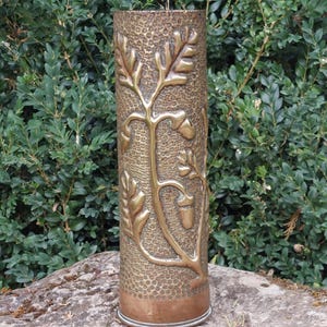 May include: A tall, cylindrical brass vase featuring a textured, hammered background surface. The exterior is decorated with a raised, embossed relief design of oak leaves and acorns that wrap around the body of the vessel in a vertical pattern.