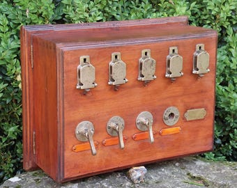 Può includere: Un'antica scatola di controllo elettrica in legno con elementi in ottone. La scatola presenta sei interruttori a levetta in ottone nella parte superiore e quattro interruttori a leva in basso. Sono visibili anche piccole targhette in ottone e dettagli arancioni.