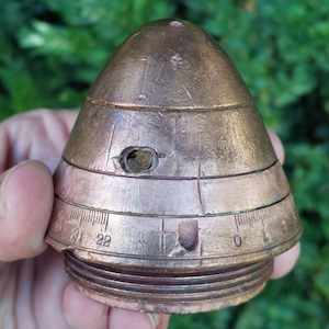 Puede incluir: Objeto cónico de color cobre con base roscada y bandas horizontales. Presenta un pequeño orificio y una escala numerada del 0 al 22. La superficie muestra signos de desgaste, lo que sugiere que es un artículo antiguo o vintage.