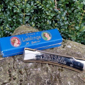 May include: A silver harmonica with the words "Unsere Liebling M. Hohner" engraved on it. The harmonica is in its original blue box with a picture of a woman on the front.