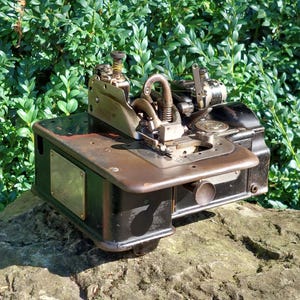 Könnte beinhalten: Eine antike schwarze Nähmaschine mit einem Metallgehäuse und silbernen Akzenten. Die Maschine hat eine große Metallplatte mit einem kleinen Typenschild an der Seite. Die Maschine steht auf einem Stein.