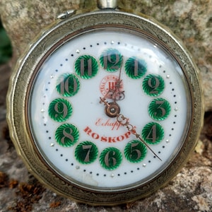 May include: A vintage pocket watch with an ornate metallic casing and a white dial. The dial features twelve circular green enamel markers with silver numerals, a central red logo, and the text "Echappement ROSKOP" in red lettering.
