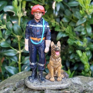 Peut inclure: Figurine d'un pompier en uniforme bleu marine, casque rouge et sangle bleu clair, à côté d'un berger allemand marron et beige. La figurine est sur une base en pierre, sur fond de feuillage vert.