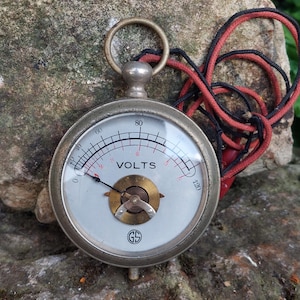 Puede incluir: Un voltímetro de bolsillo vintage de plata con un cable negro y rojo. La cara del medidor es blanca con marcas negras y una aguja roja. El medidor lee "VOLTS" y tiene un rango de 0 a 120. El medidor tiene un logotipo GS.