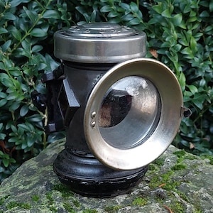 Puede incluir: Una linterna antigua de metal negro y plateado con una lente de vidrio grande y redonda. La linterna tiene una parte superior plateada circular y un borde plateado alrededor de la lente. La linterna está sobre una roca cubierta de musgo.