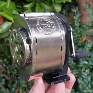 Peut inclure: Un taille-crayon argenté vintage avec une poignée et une base noires. Le taille-crayon présente un design circulaire avec plusieurs trous et le texte "Multi Hole Sharpener". Le taille-crayon est tenu dans une main sur un fond de feuillage vert.