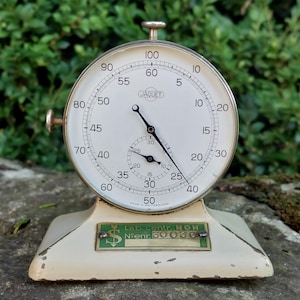 Könnte beinhalten: Eine antike Jaquet Stoppuhr mit cremefarbenem Zifferblatt und schwarzen Ziffern. Das Metallgehäuse steht auf einem cremefarbenen Sockel mit grün-goldener Beschriftung. Ein klassisches Zeitmessgerät, hergestellt in der Schweiz.