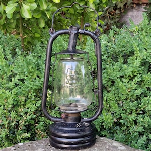 Pode incluir: Uma lanterna preta vintage com um globo de vidro transparente, com uma alça e uma base. O globo de vidro tem um logotipo. A lanterna está colocada sobre um fundo de folhagem verde. A altura da lanterna é de aproximadamente 30 cm.
