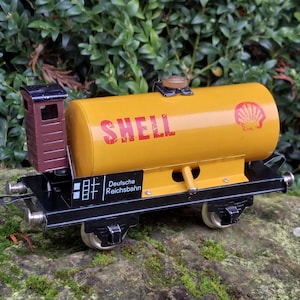 Könnte beinhalten: Ein alter Spielzeug-Eisenbahnwagen mit einem gelben Tank mit der roten Aufschrift "SHELL". Der Wagen hat eine braune Kabine und ein schwarzes Fahrgestell mit dem Text "Deutsche Reichsbahn". Der Wagen steht auf einer Steinoberfläche.