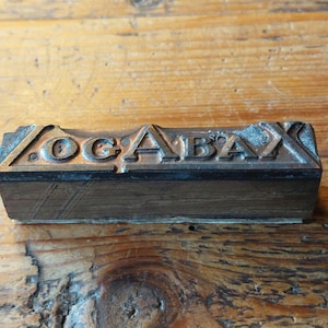 May include: A close-up of a metal printing block with the word "OG'ABAX" embossed on it. The block is resting on a wooden surface.