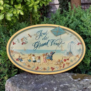 Peut inclure: Panneau ovale en bois représentant une scène de plage avec le texte "Jour de Grand Vent". Le panneau montre des enfants jouant, un cerf-volant et une tente rayée. La bordure est jaune et bleue, avec un fond bleu clair.